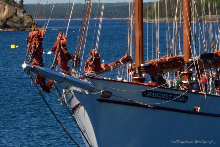 Two sailboats docked with brown sails, blue water and forested shore in the background.