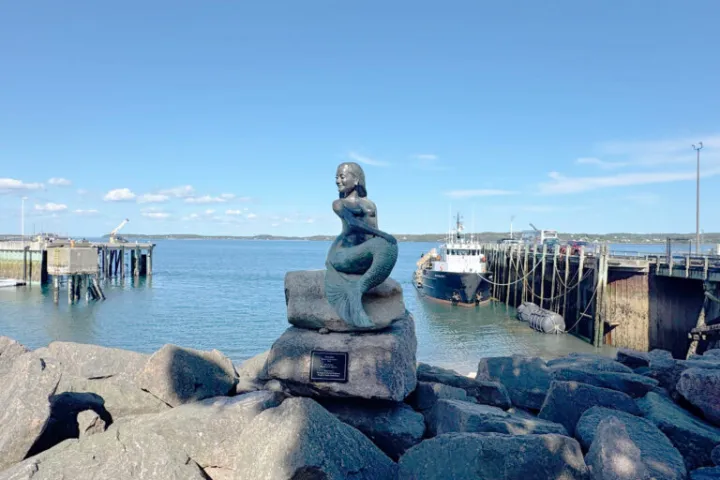Mermaid statue on rocks by a harbor with boats and clear sky.