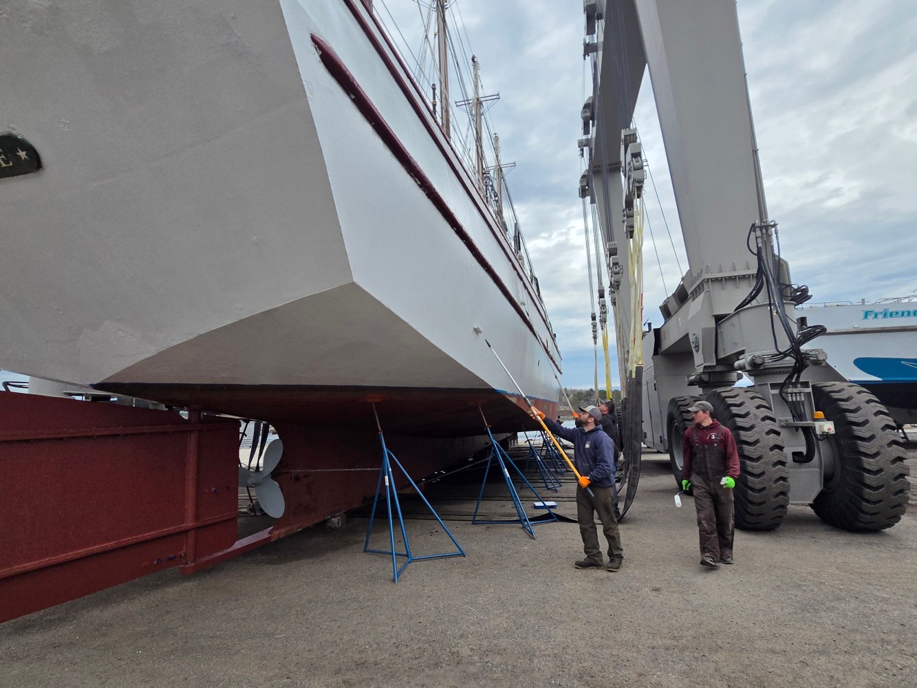 People inspecting large boat on supports with crane nearby.