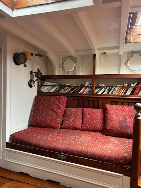 Cozy boat cabin with red cushioned bench, wooden shelves, and porthole windows.