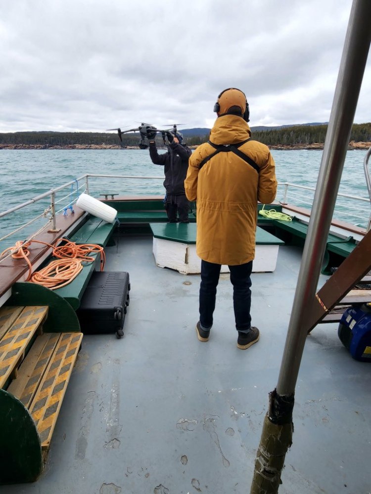 Two people on a boat deck, one holding a drone, with water and hills in the background.