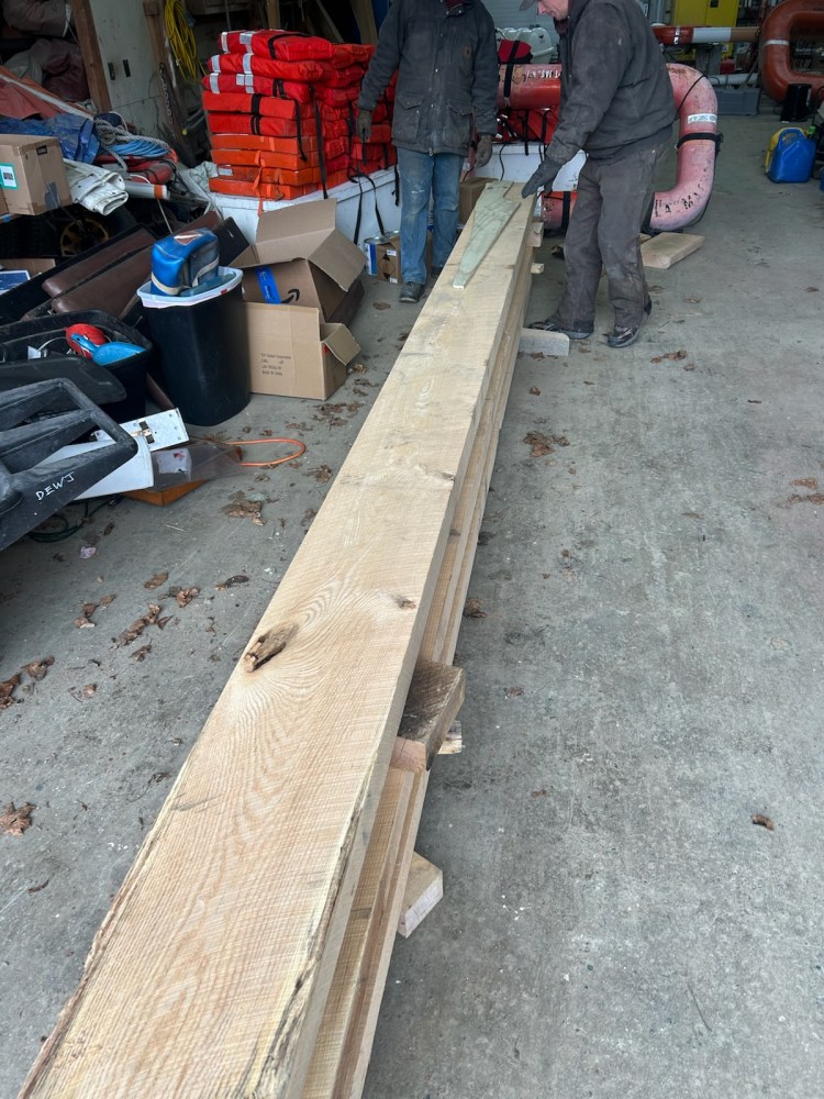 Two people handling long wooden planks in a cluttered workshop.