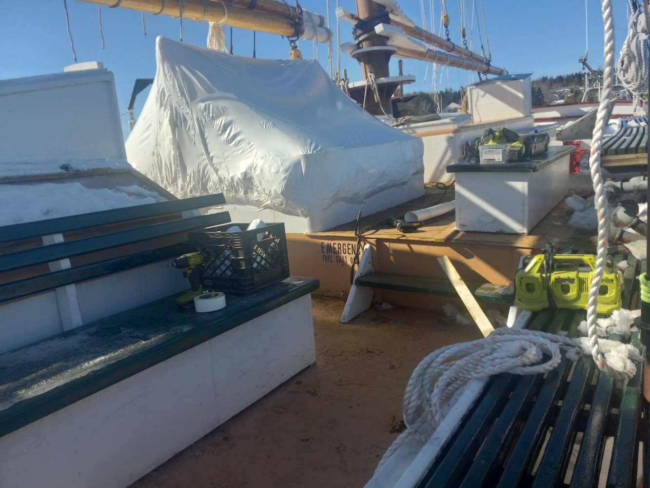 Boat deck with covered equipment and benches, ropes, and clear sky.