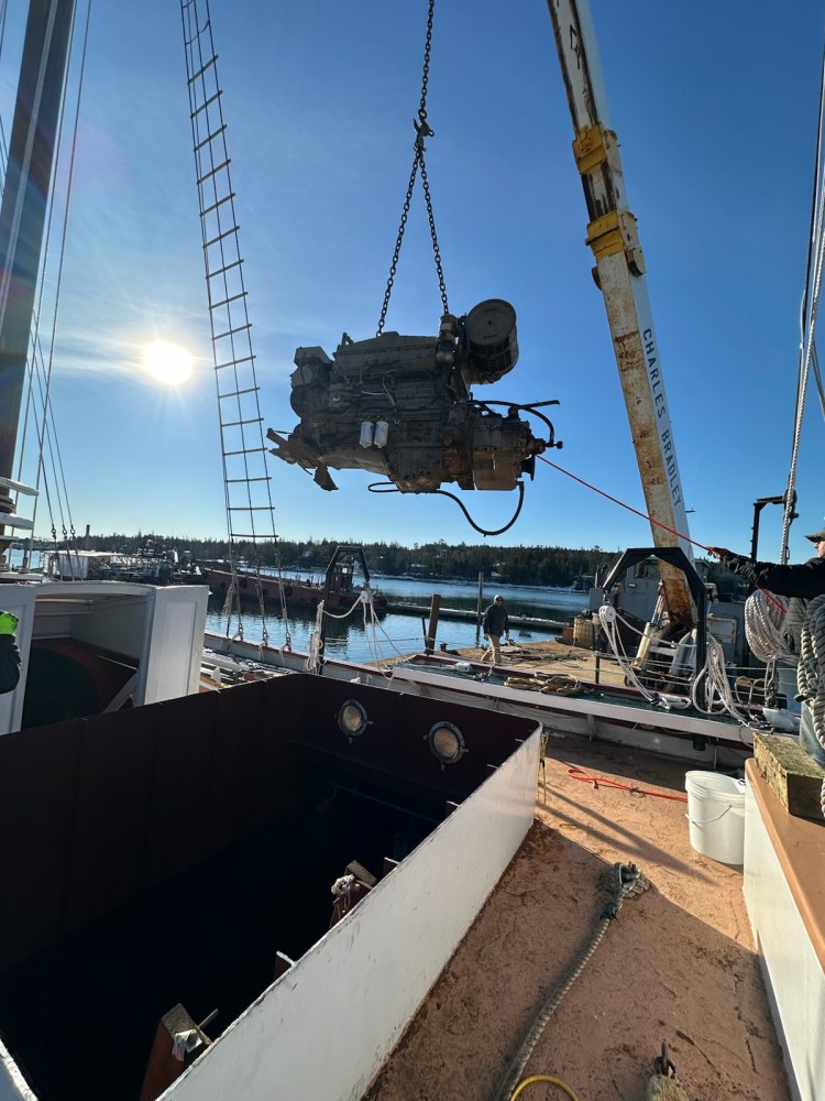 Crane lifting large engine part from dock under a clear blue sky.