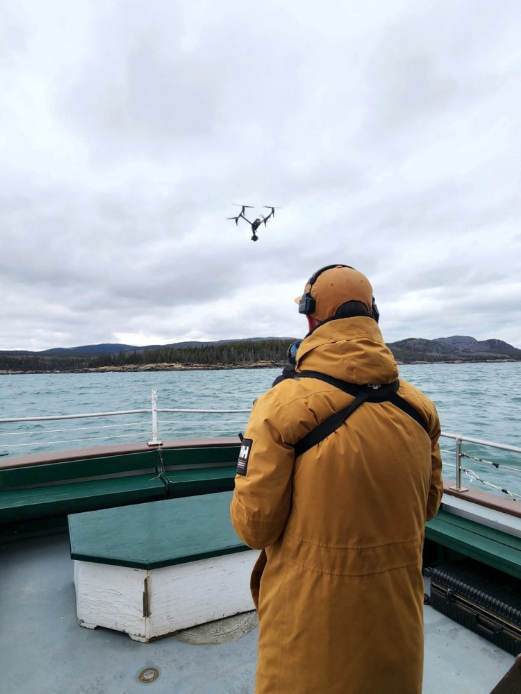 Person in yellow coat on boat operates a drone over water.